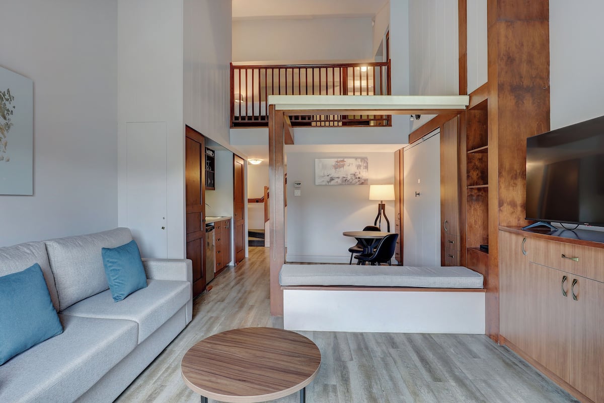 A modern living room with a sofa, coffee table, and a TV. There is a staircase leading to an upper level.