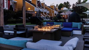 Un espacio de estar al aire libre con mobiliario de mimbre, una chimenea central y edificios residenciales de fondo.