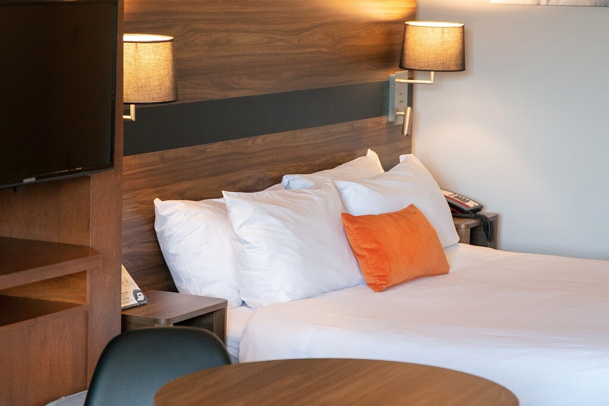 A hotel room with a bed, white bedding, an orange pillow, a TV, and two wall-mounted lamps.