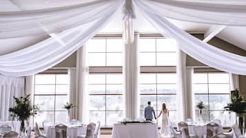 Una recepción de boda con vistas a un paisaje nevado a través de amplias ventanas, cortinas blancas con drapeados y mesas dispuestas para los invitados.