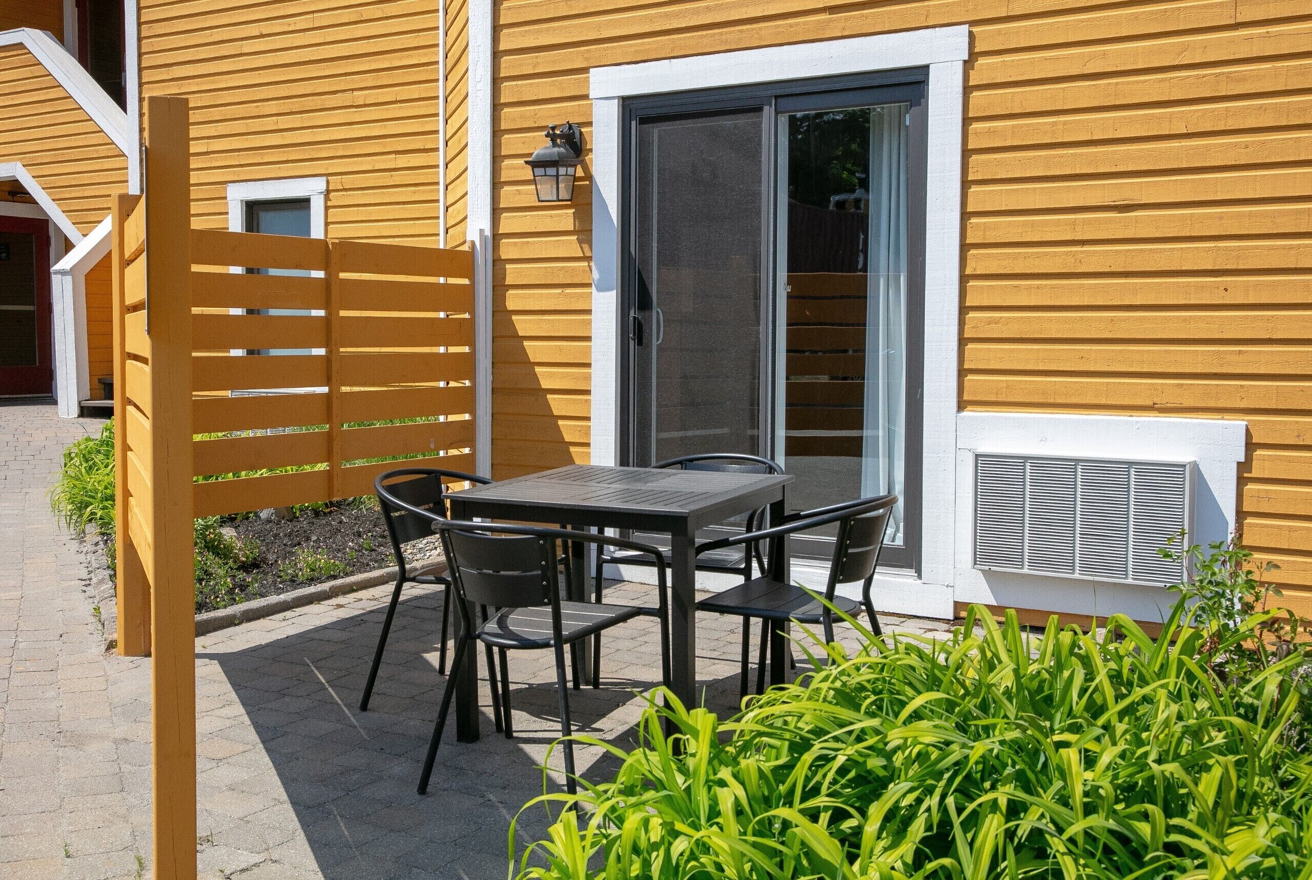An outdoor patio with a table and chairs, a wooden fence, and a sliding glass door to a room.
