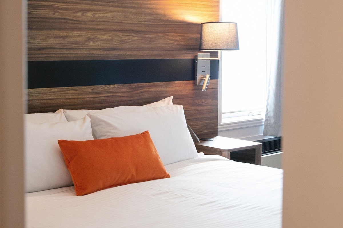 A hotel room with a wooden headboard, a white bed with an orange pillow, a bedside table, and a lamp.
