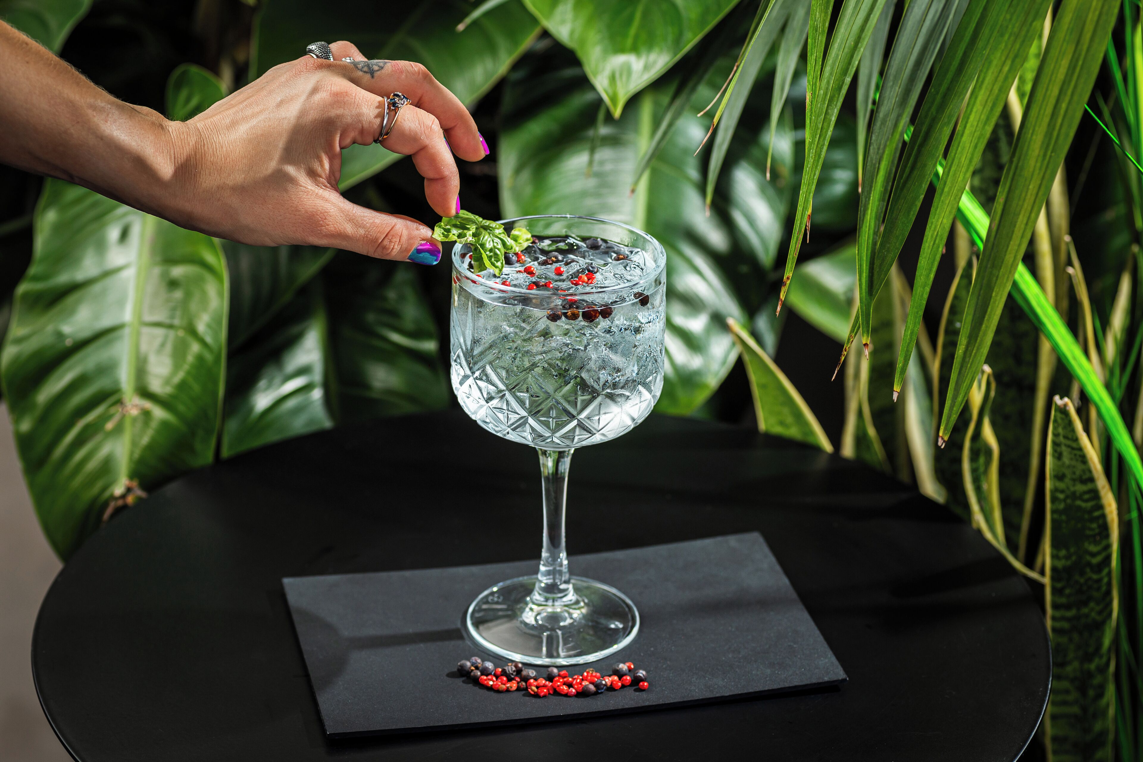 A hand placing a mint leaf into a glass with ice and red peppercorns.