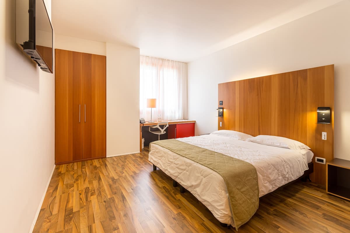 A hotel room with a wooden floor, a bed with a quilt, a desk with a chair, a red cabinet, and a wall-mounted TV.