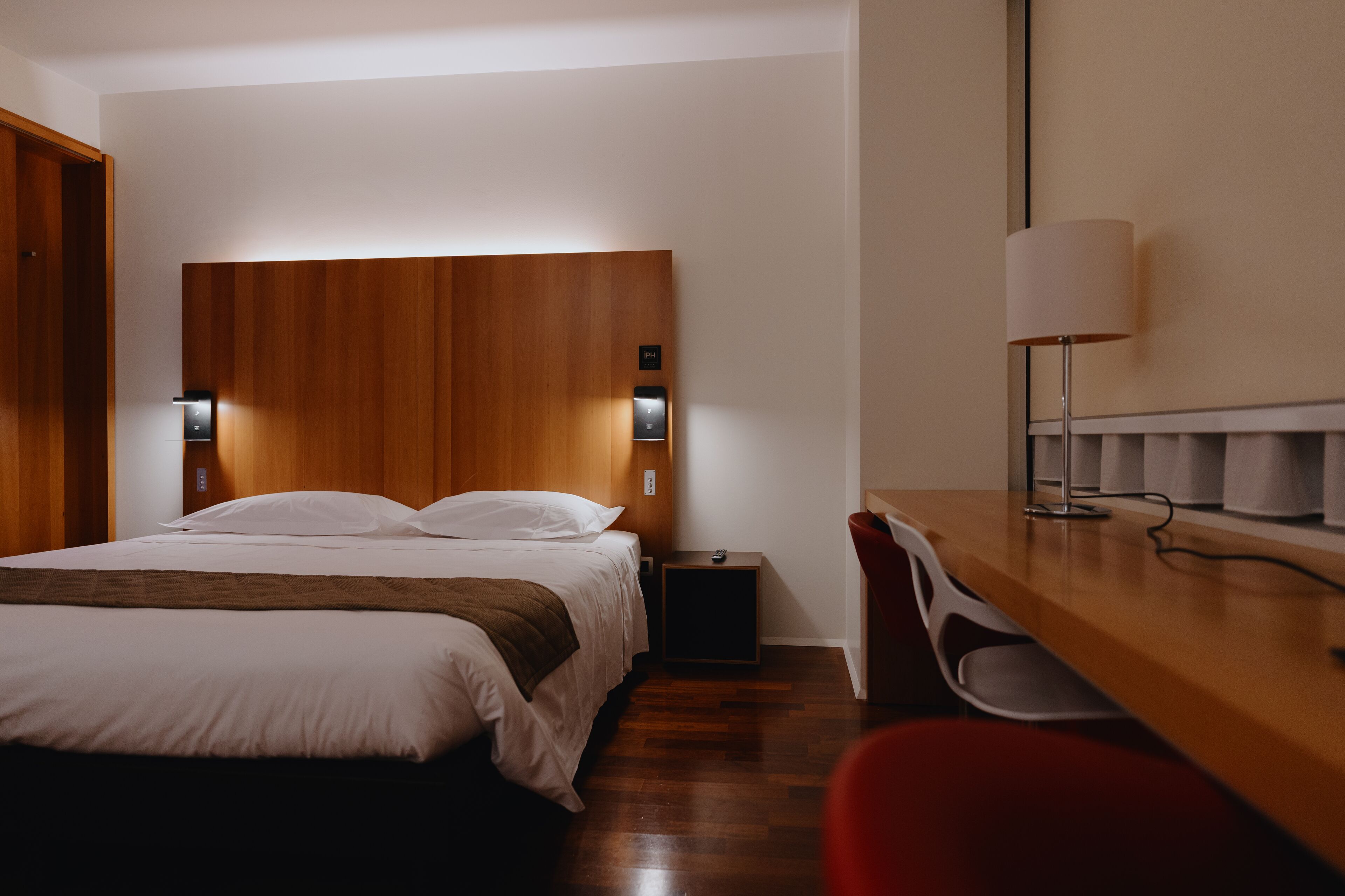 Een moderne hotelkamer met een houten hoofdeinde, een bed met witte lakens, een nachtkastje met een lamp en een rode fauteuil.