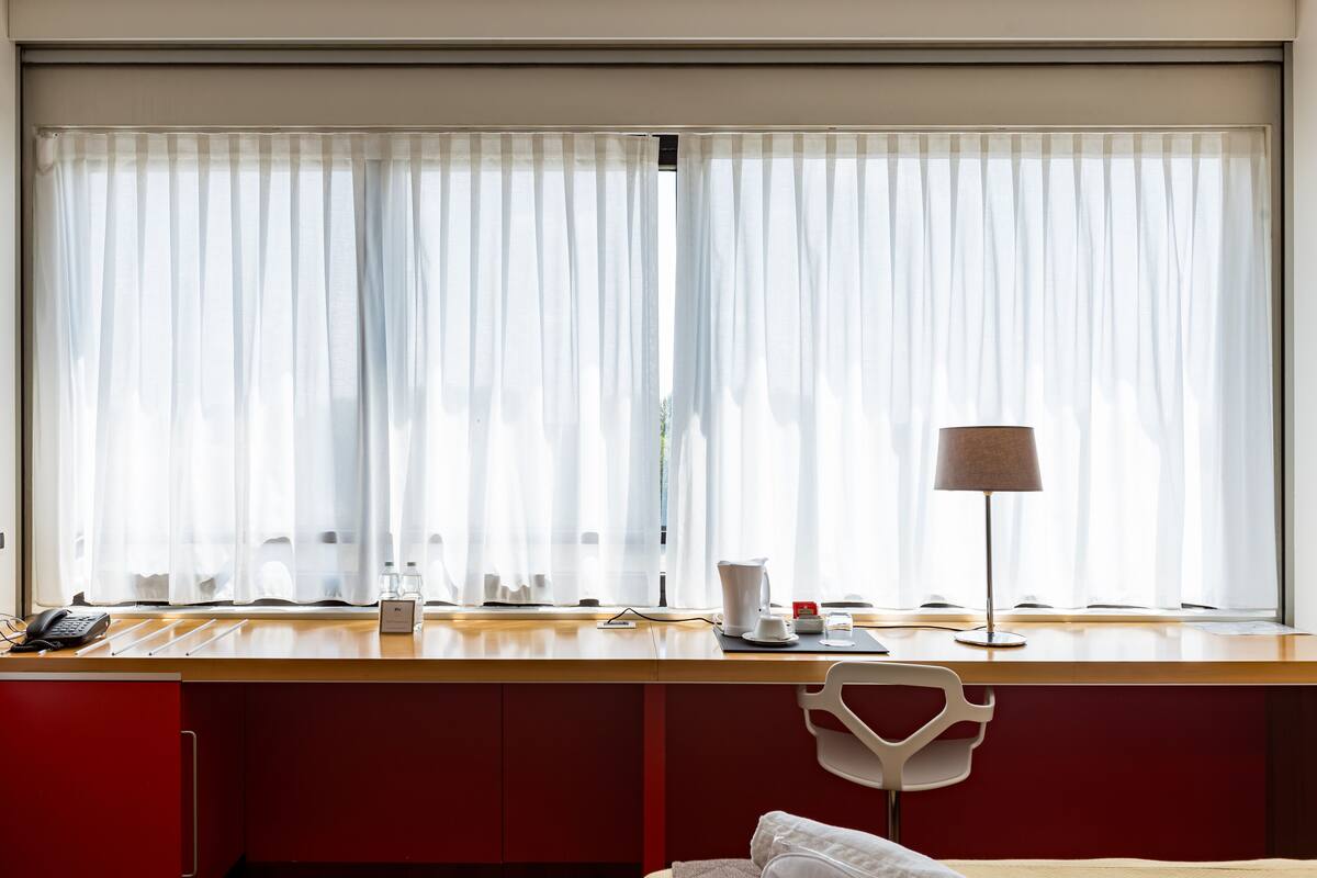 A hotel room with a wooden desk, a white chair, a bed with white bedding, a lamp, and a window with sheer curtains.