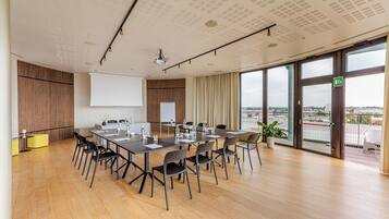 Una sala de conferencias moderna con una mesa grande, sillas, una pantalla de proyector y vistas a la ciudad a través de amplias ventanas.