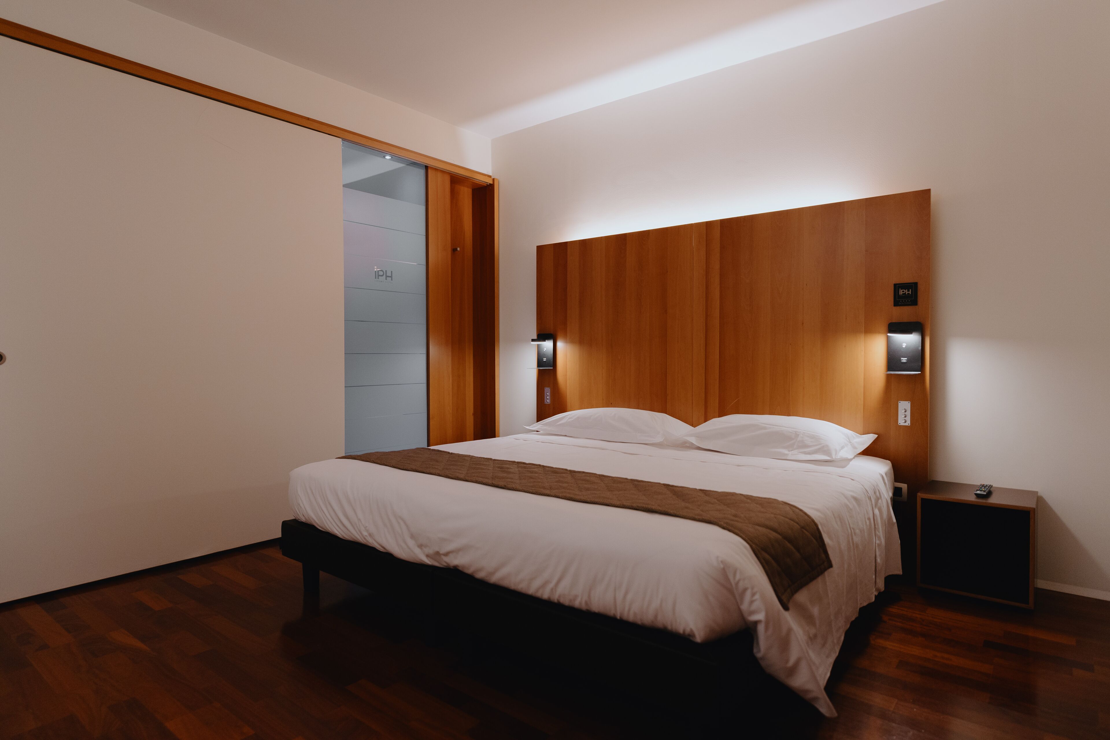 A modern bedroom with a wooden headboard, a bedside table with a remote, and a sliding door to the left.