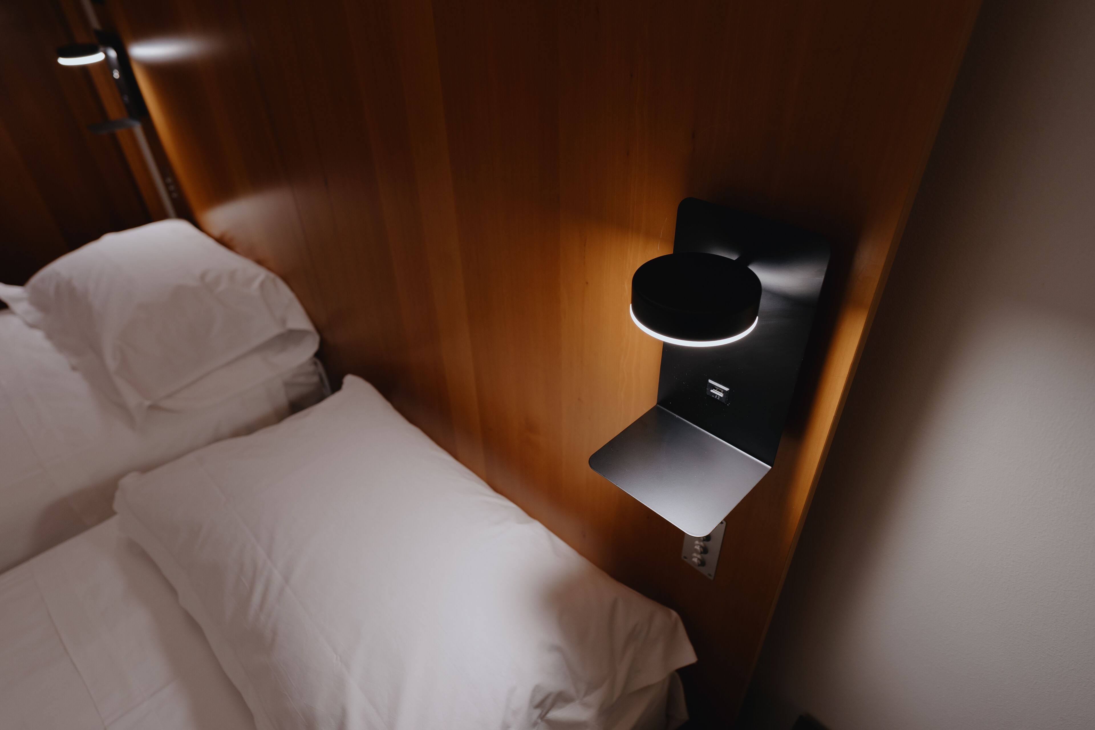 Une chambre d’hôtel moderne avec un mur en bois, un lit recouvert de draps blancs et un luminaire fixé au mur.