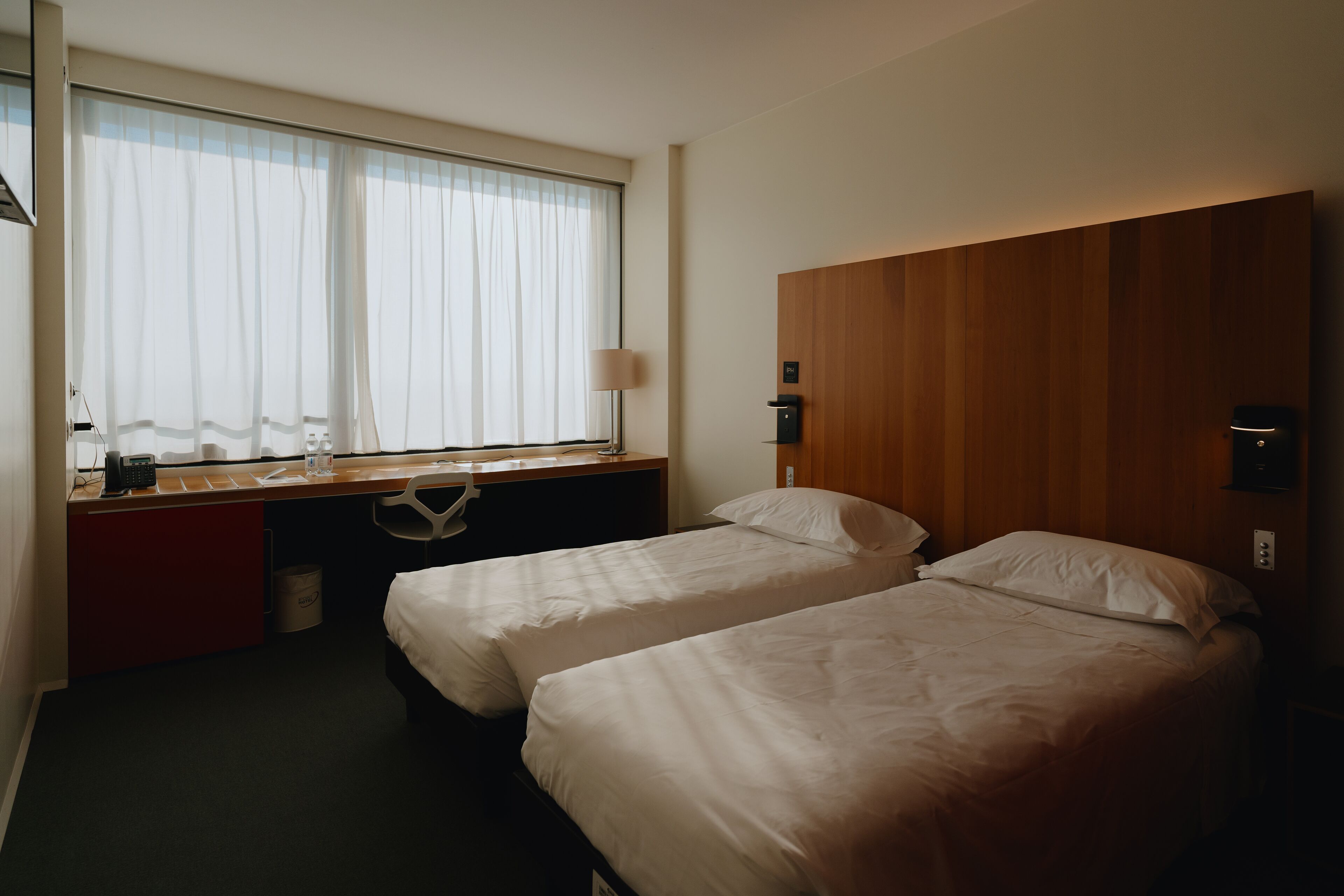 Camera d'albergo con due letti singoli, una testiera in legno, una scrivania con lampada e una finestra con tende leggere.