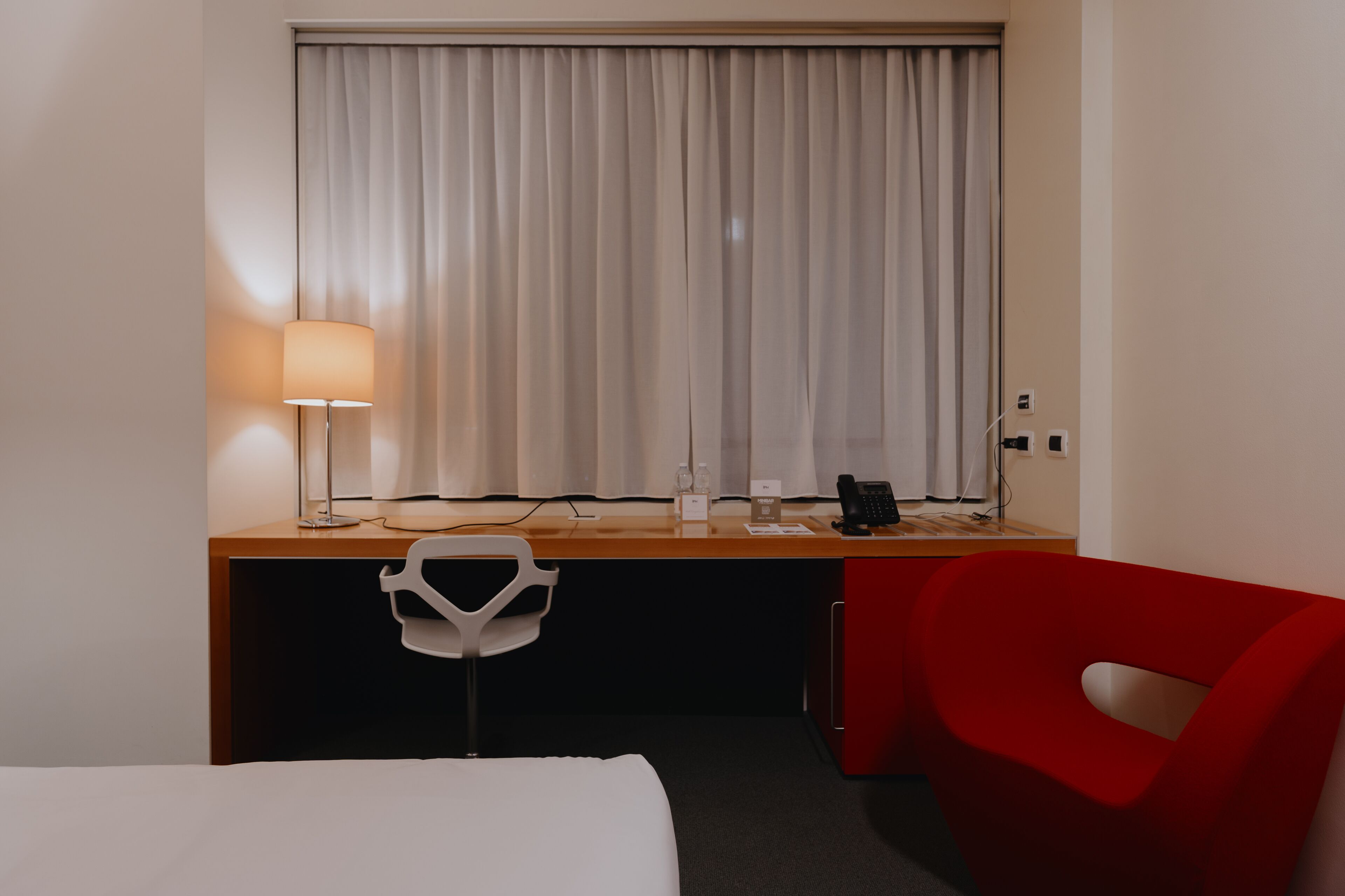 Een hotelkamer met een bed, een bureau met lamp, een rode stoel en een raam met gordijnen.