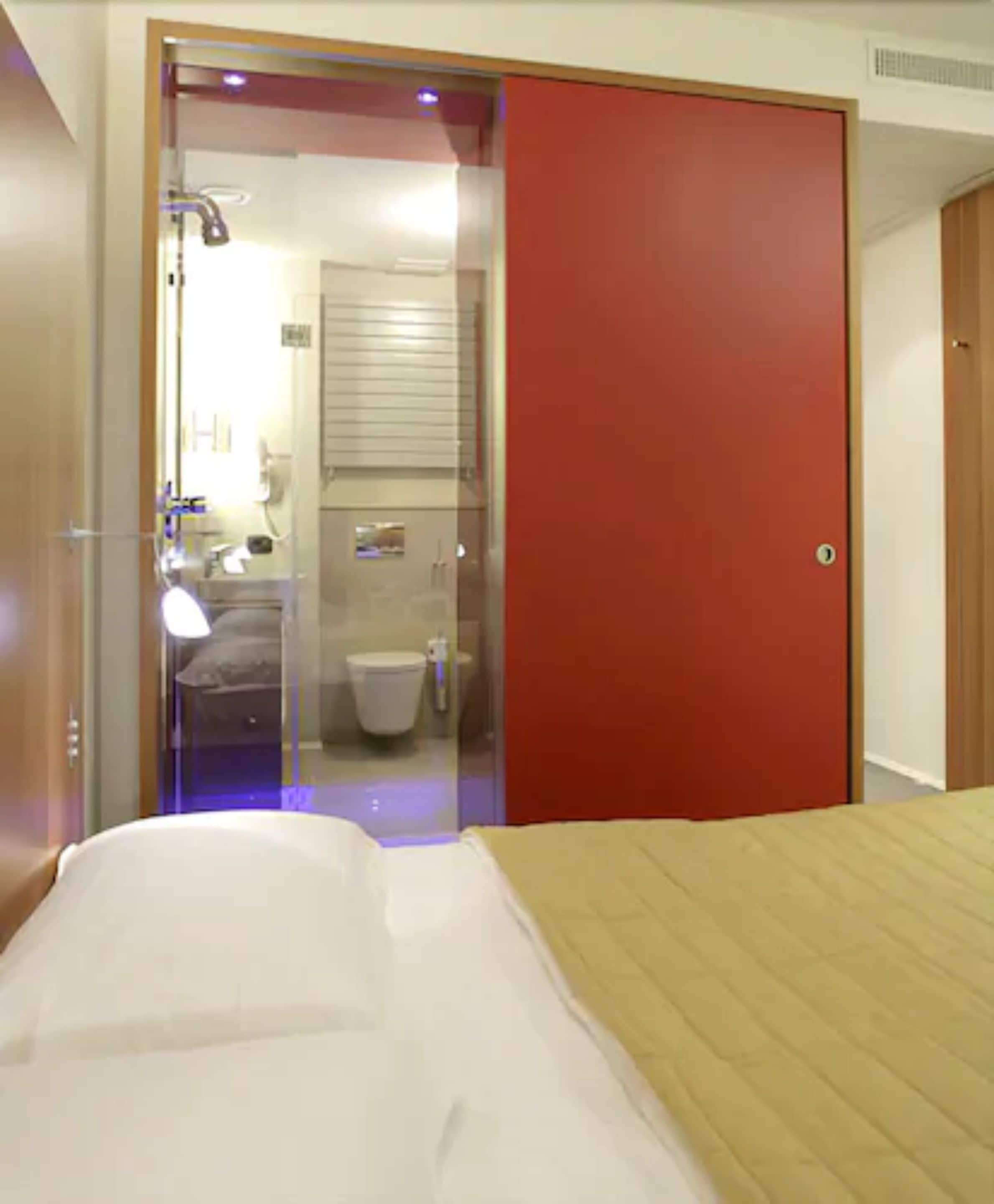 A hotel room with a bed, a red door, and a bathroom visible through a glass door.