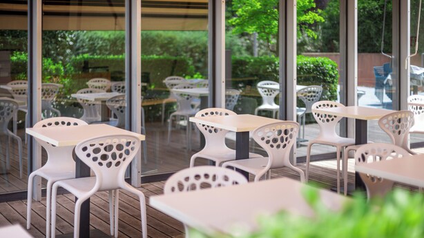 Un espace extérieur aménagé avec des tables et des chaises blanches, de grandes fenêtres et de la verdure visible à travers le verre.