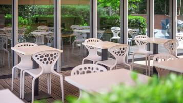 Un espace extérieur aménagé avec des tables et des chaises blanches, de grandes fenêtres et de la verdure visible à travers le verre.