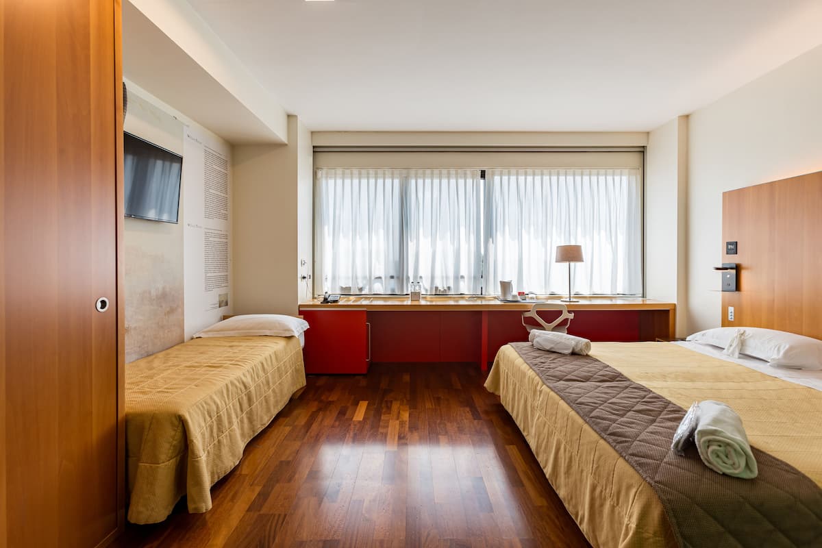 A hotel room with two beds, a desk, a window with curtains, and a wooden floor.