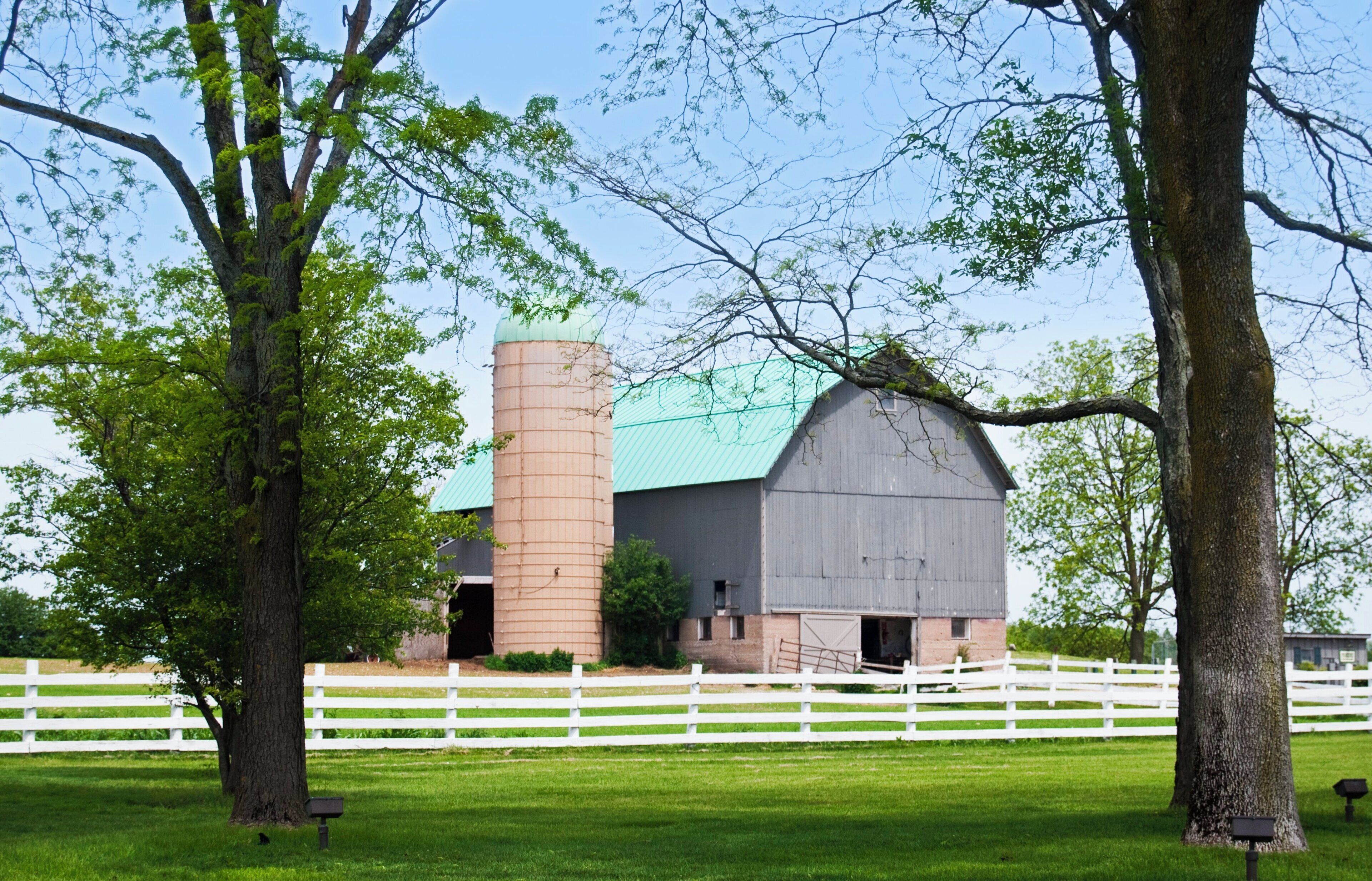 A barn with a silo, surrounded by a white fence, trees, and a clear sky.