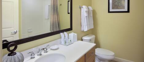 A bathroom with a white sink, mirror, and toilet. There are towels hanging on the wall and a framed picture above the toilet.
