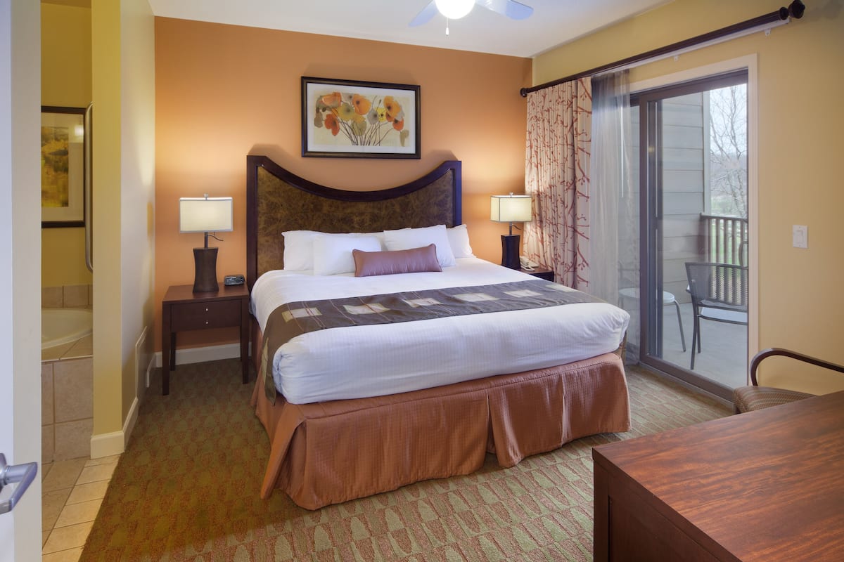 A hotel room with a large bed, two bedside tables with lamps, a balcony with a chair, and a painting above the bed.