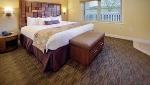 A hotel room with a large bed, a wooden headboard, a bench at the foot of the bed, a nightstand, and a window with blinds. - Holiday Inn Club Vacations at Lake Geneva Resort by IHG (Lake Geneva)