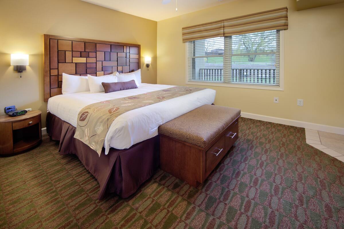 A hotel room with a large bed, a wooden headboard, a bench at the foot of the bed, a nightstand, and a window with blinds.