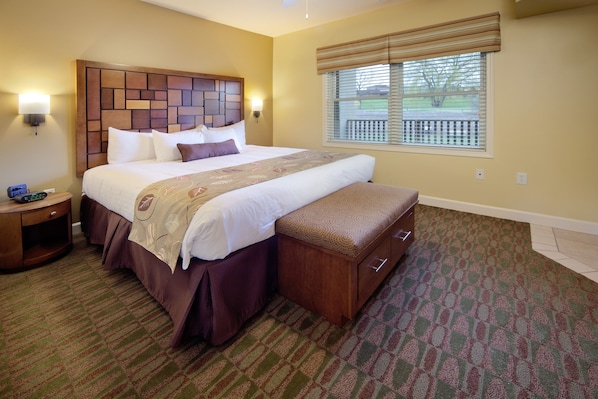 Quarto de hotel com uma cama grande, cabeceira de madeira, um banco no pé da cama, mesa de cabeceira e uma janela com persianas.