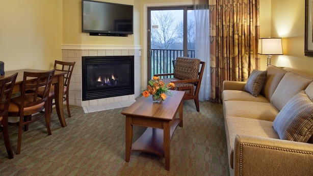 A living room with a fireplace, a television mounted above it, a sofa, a coffee table, and a view through a large window. - Holiday Inn Club Vacations at Lake Geneva Resort by IHG (Lake Geneva)