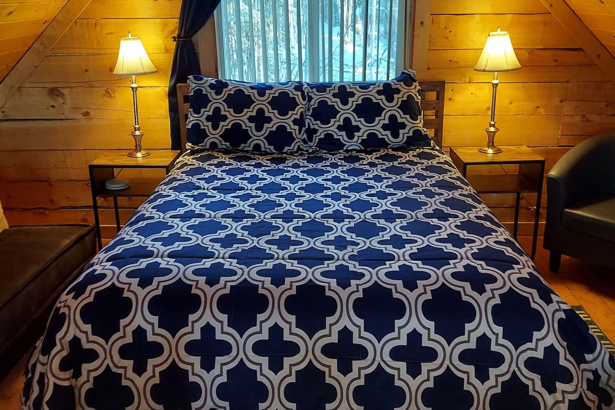 Une chambre confortable située sous les combles, avec un plafond en pente en bois, un grand lit recouvert d’une courtepointe à motifs, deux lampes de chevet et une fenêtre avec des rideaux.