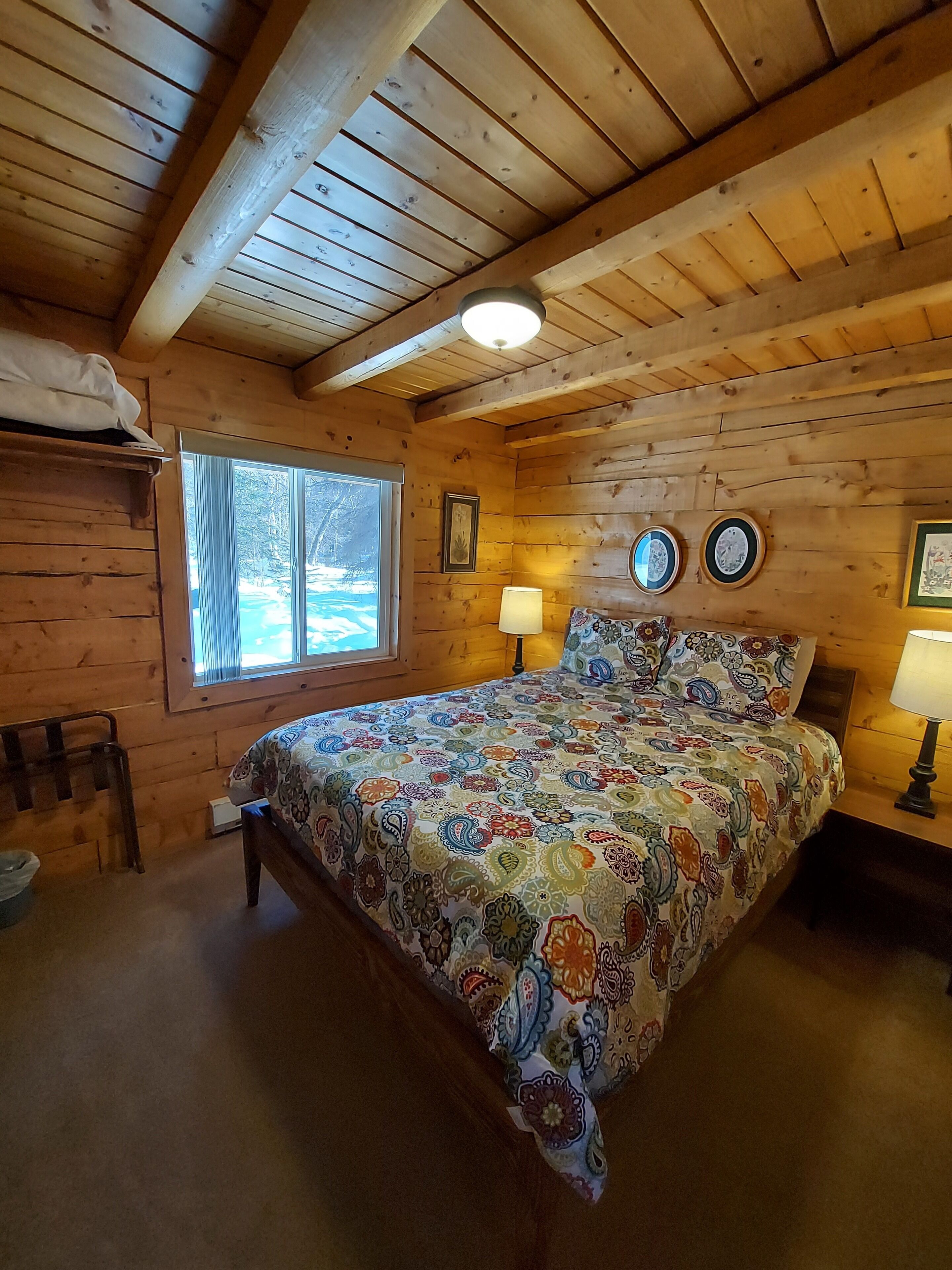 A wooden cabin bedroom with a bed, two bedside lamps, a window, and a wooden ceiling.
