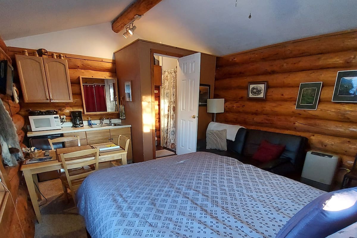 Une pièce chaleureuse aménagée dans un chalet en rondins, avec un lit, un bureau équipé d’un ordinateur, un ventilateur de plafond et des murs en bois.