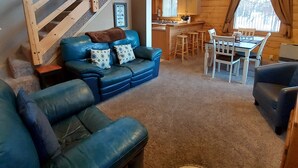 A cozy cabin interior with a blue leather sofa, a dining table with chairs, a kitchen area, and a wooden staircase. - Hatcher Pass Cabins (Palmer)