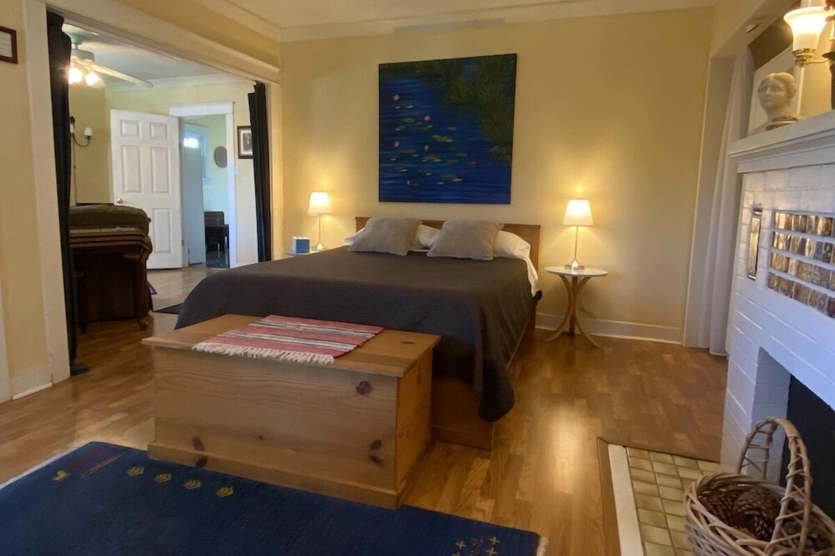 A hotel room with a bed, two bedside lamps, a painting on the wall, a wooden chest, and a fireplace.