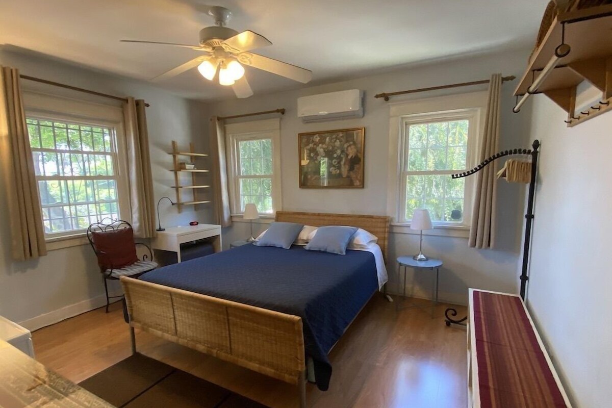 A bedroom with a bed, a desk, a chair, a ceiling fan, and a window.