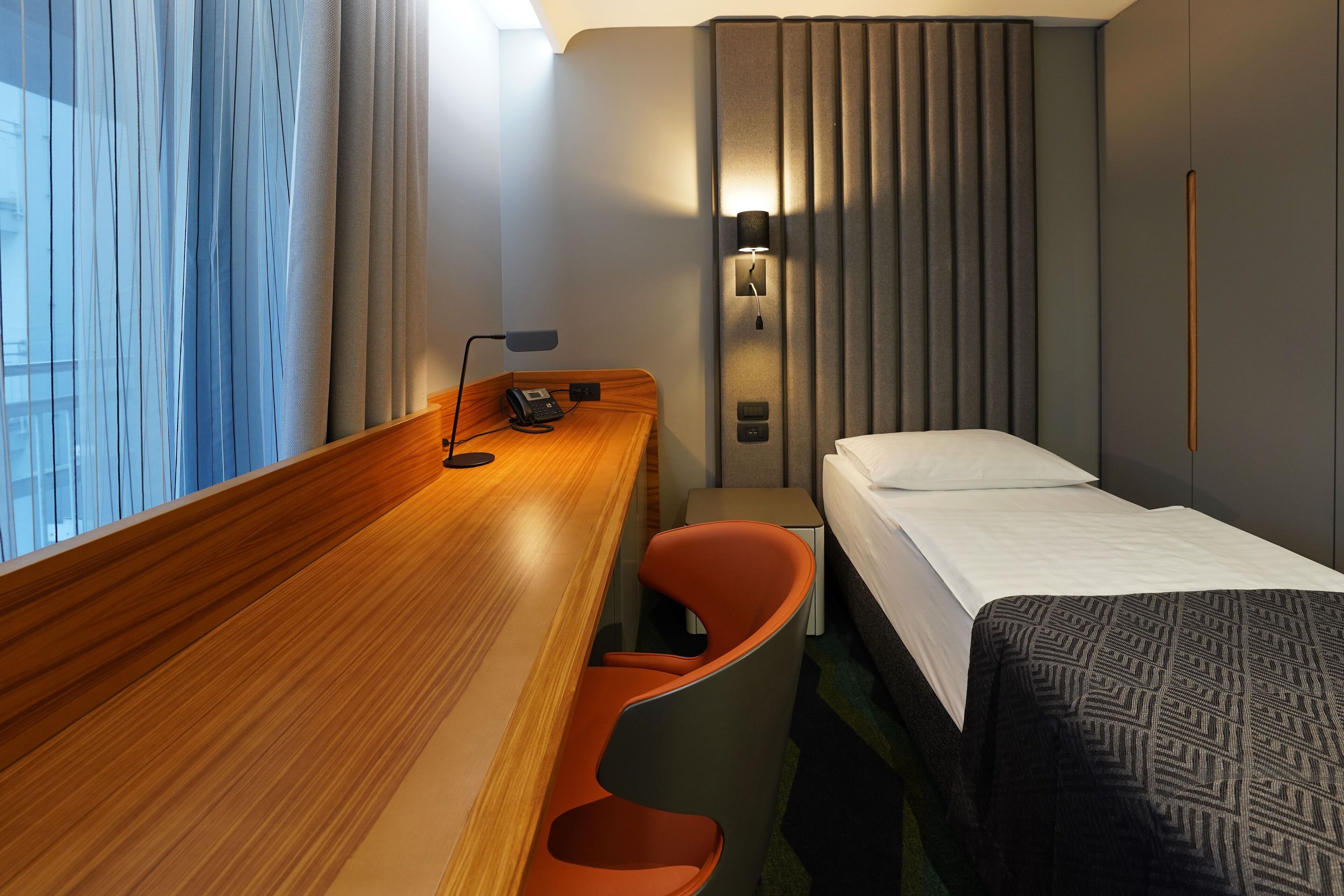 Une chambre d’hôtel avec un bureau en bois, un lit, une chaise, une table de chevet, une lampe et une fenêtre avec des rideaux.