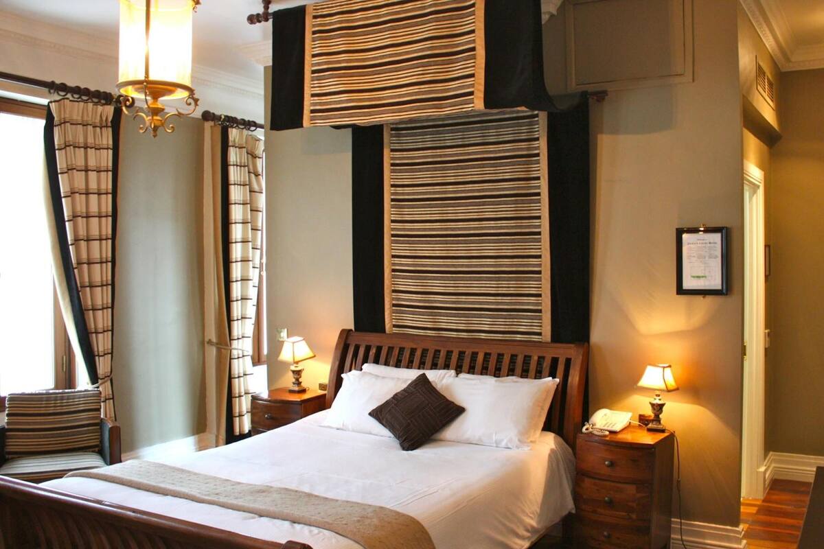 Una cama bien tendida con un cabecero de madera, dos lámparas de noche y un certificado enmarcado en la pared.
