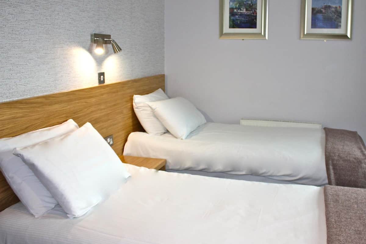 Habitación de hotel con dos camas, cabecera de madera, ropa de cama blanca, una mesita de noche y dos cuadros en la pared.