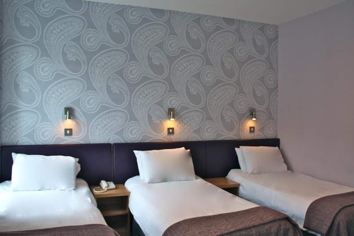 Habitación de hotel con tres camas, cada una con ropa blanca y mantas marrones. Mesitas de noche con lámparas y teléfono. La pared presenta un estampado paisley.