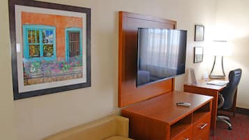 Habitación de hotel con un televisor de pantalla plana grande montado en la pared, un escritorio de madera con una silla, un banco acolchado y un cuadro enmarcado de una casa.
