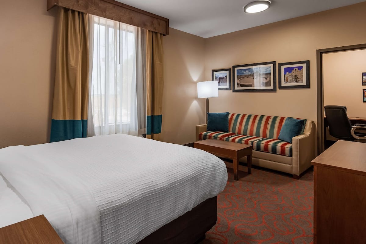 Quarto de hotel com uma cama grande, um sofá pequeno, uma escrivaninha e uma cadeira. Há duas luminárias, uma janela com cortinas e quadros nas paredes.