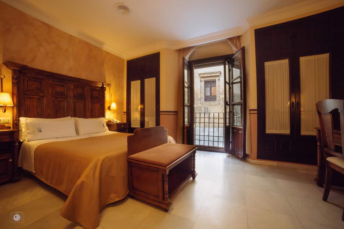Ein Hotelzimmer mit einem großen Bett, Nachttischen, einer hölzernen Bank und Blick auf die Straße durch eine offene Tür.