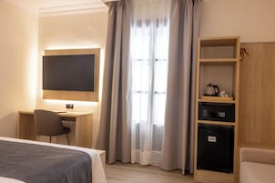 Quarto de hotel com cama, escrivaninha com cadeira, televisão, minibar e guarda-roupa.