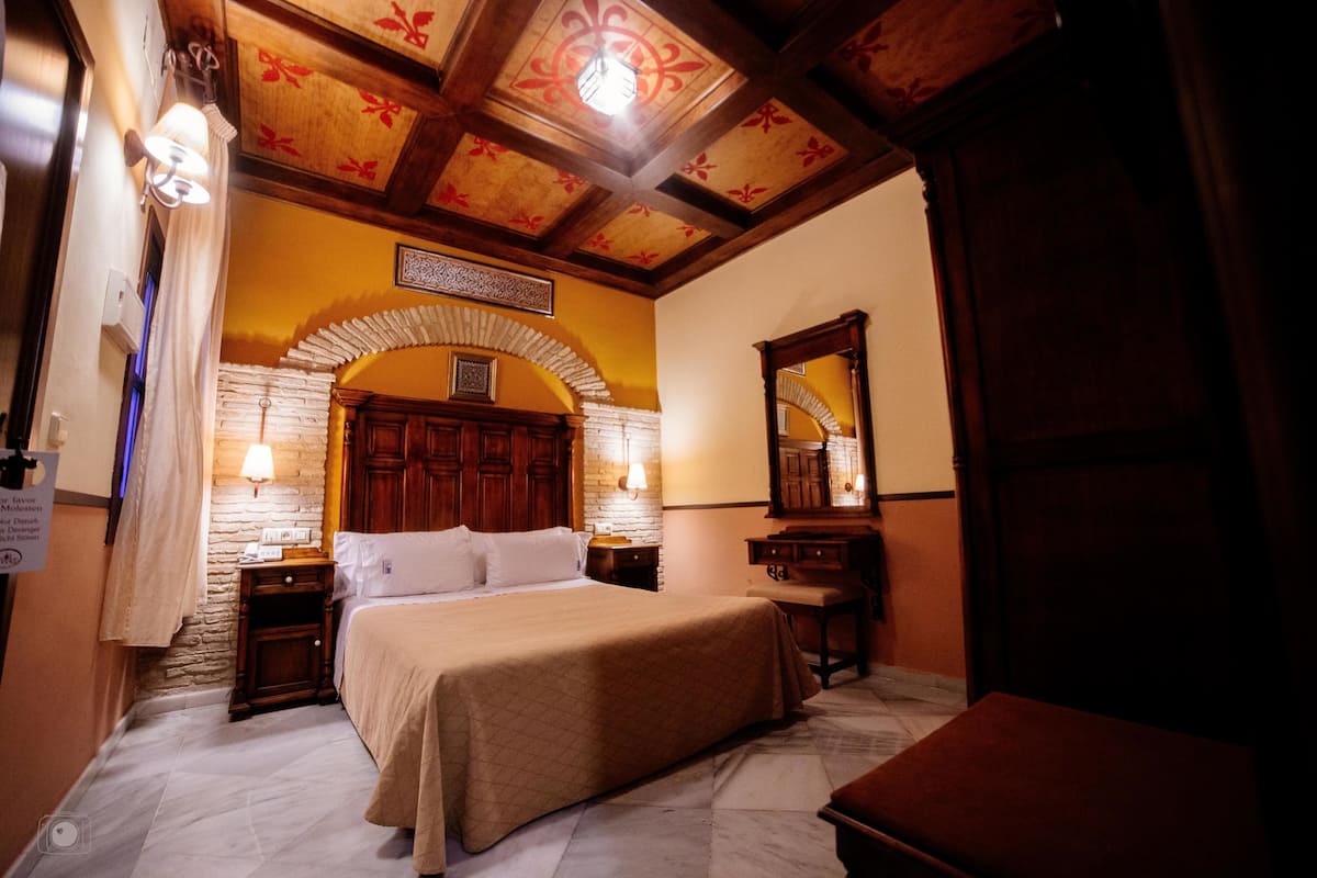 Ein Hotelzimmer mit einem Bett, einem Holz-Kopfstück, zwei Nachttischen mit Lampen, einer Bank und einem großen Spiegel.