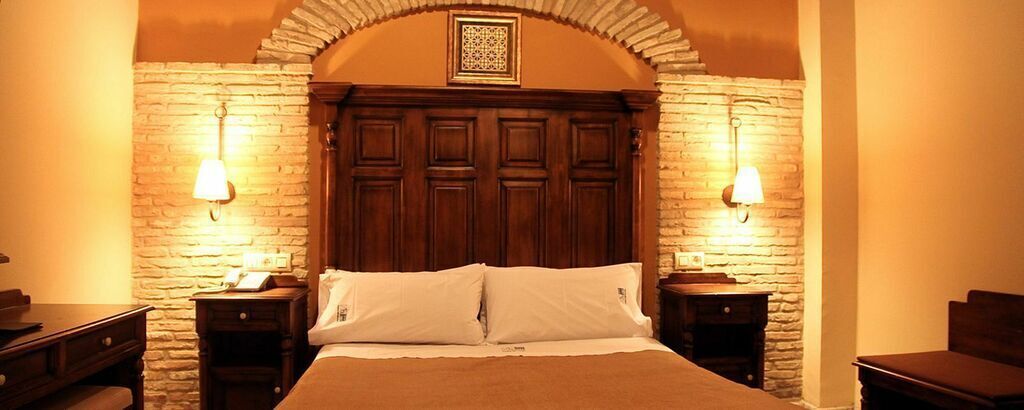 Habitación de hotel con pared de piedra, cabecero de madera, dos mesitas de noche y cama con ropa blanca.