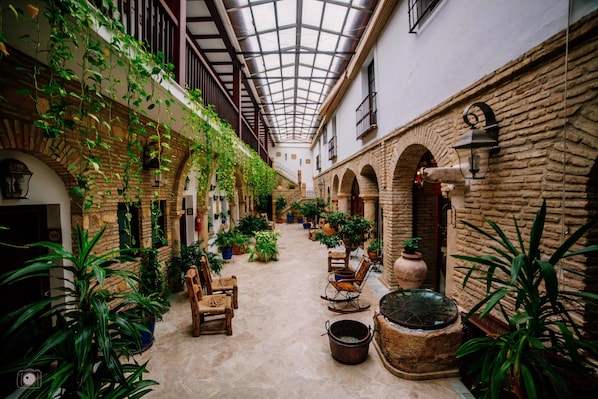 An indoor courtyard with stone walls, arches, and hanging plants. There are wooden chairs and a large pot on the ground.