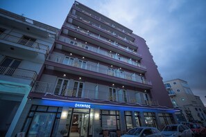 Un hotel de varios pisos con balcones, un restaurante visible llamado 'ONYMNOZ' en la planta baja y un estacionamiento al frente.
