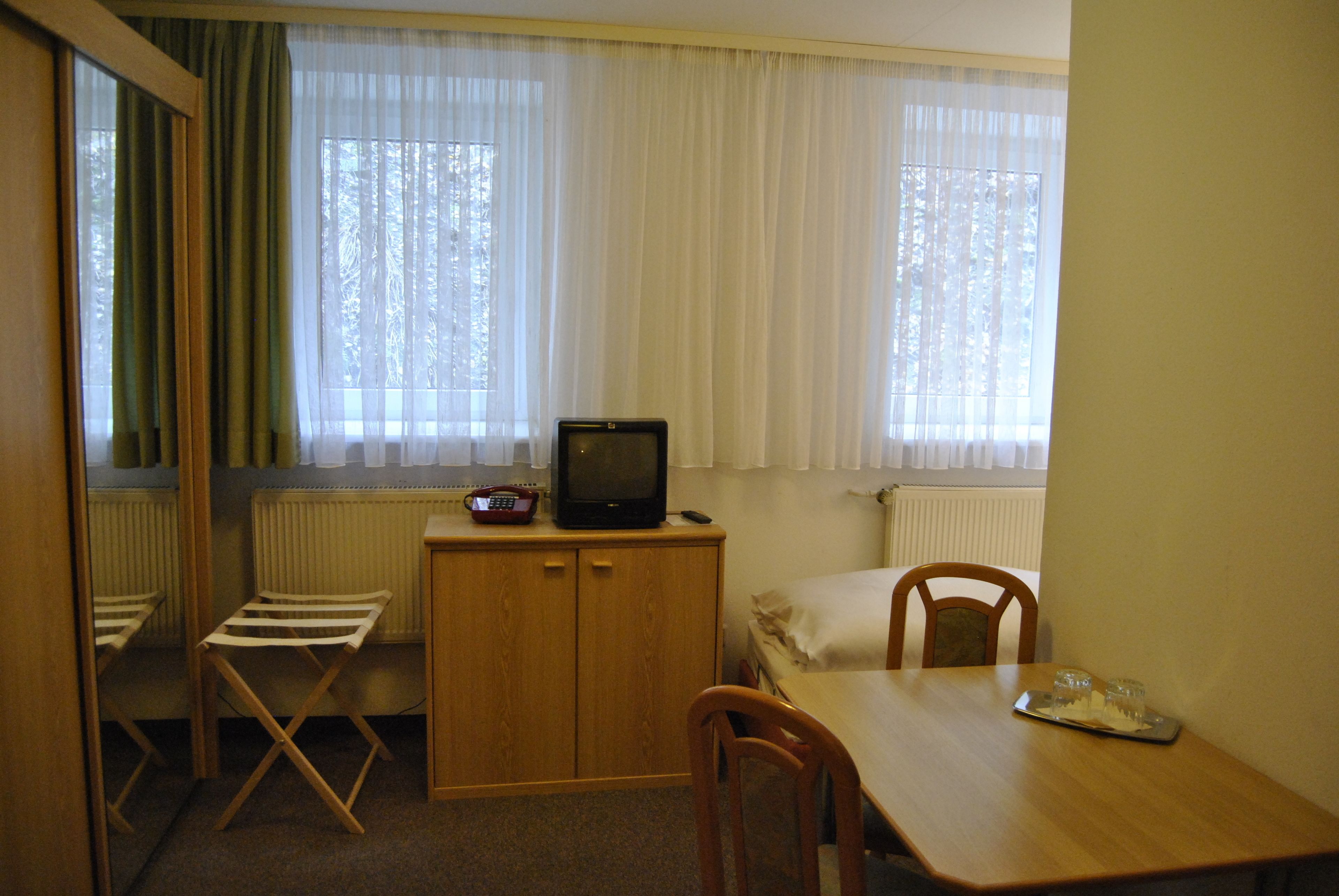 A small, compact room with a wooden table, a single bed, a television on a wooden cabinet, and a window with sheer curtains.
