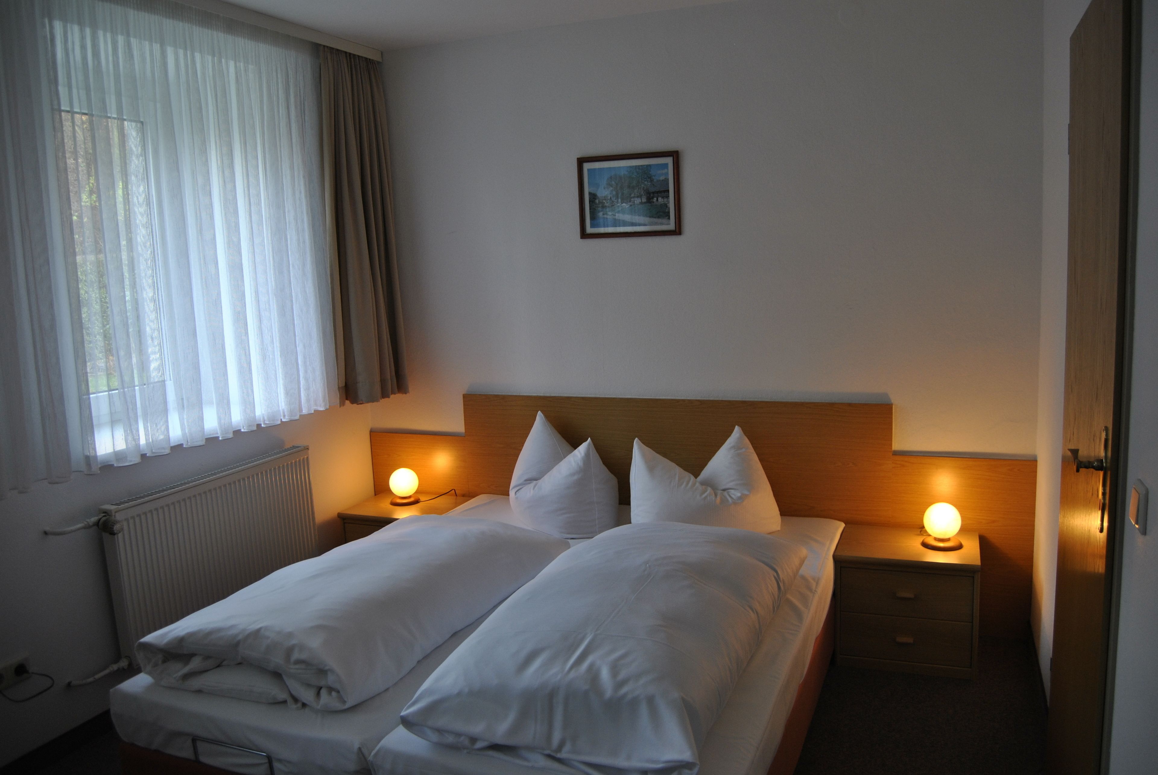 Ein Doppelbett mit weißen Bettwäsche, zwei Nachttische mit Lampen, ein gerahmtes Bild an der Wand und ein Fenster mit durchsichtigen Vorhängen.