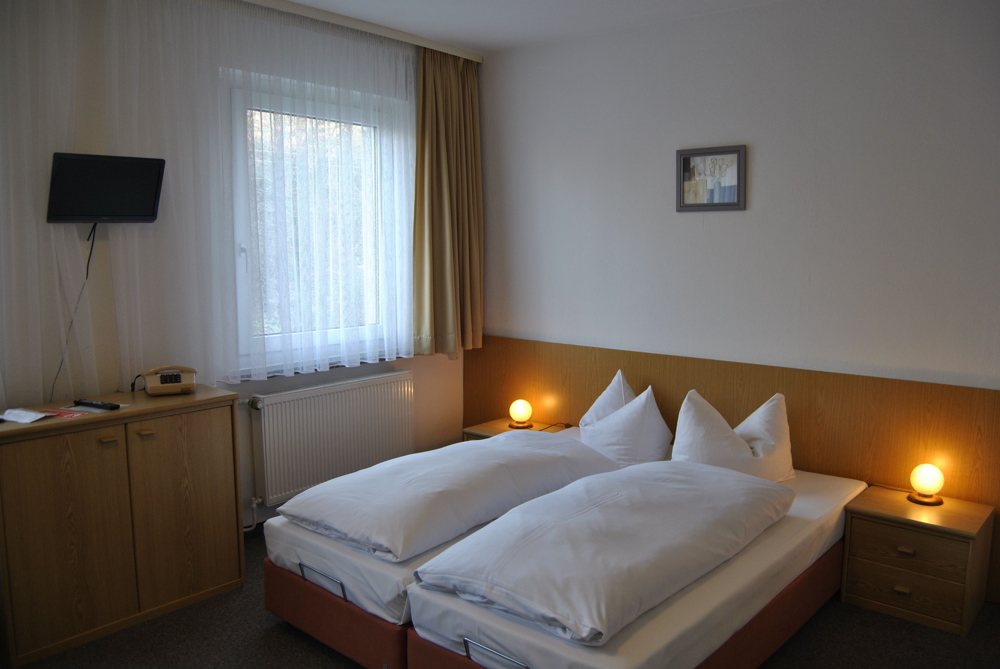 A hotel room with two beds, a television on the wall, a bedside table with a lamp, and a window with curtains.