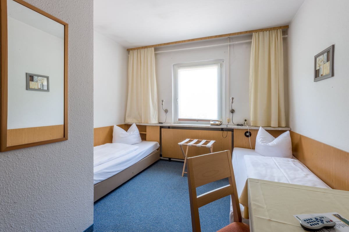 Une chambre d’hôtel avec deux lits simples, une chaise en bois, un bureau avec une chaise, une fenêtre avec des rideaux et un miroir au mur.