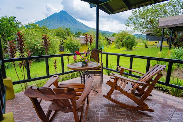 Un patio con mecedoras de madera y una mesa, con vistas a un exuberante jardín y una montaña.