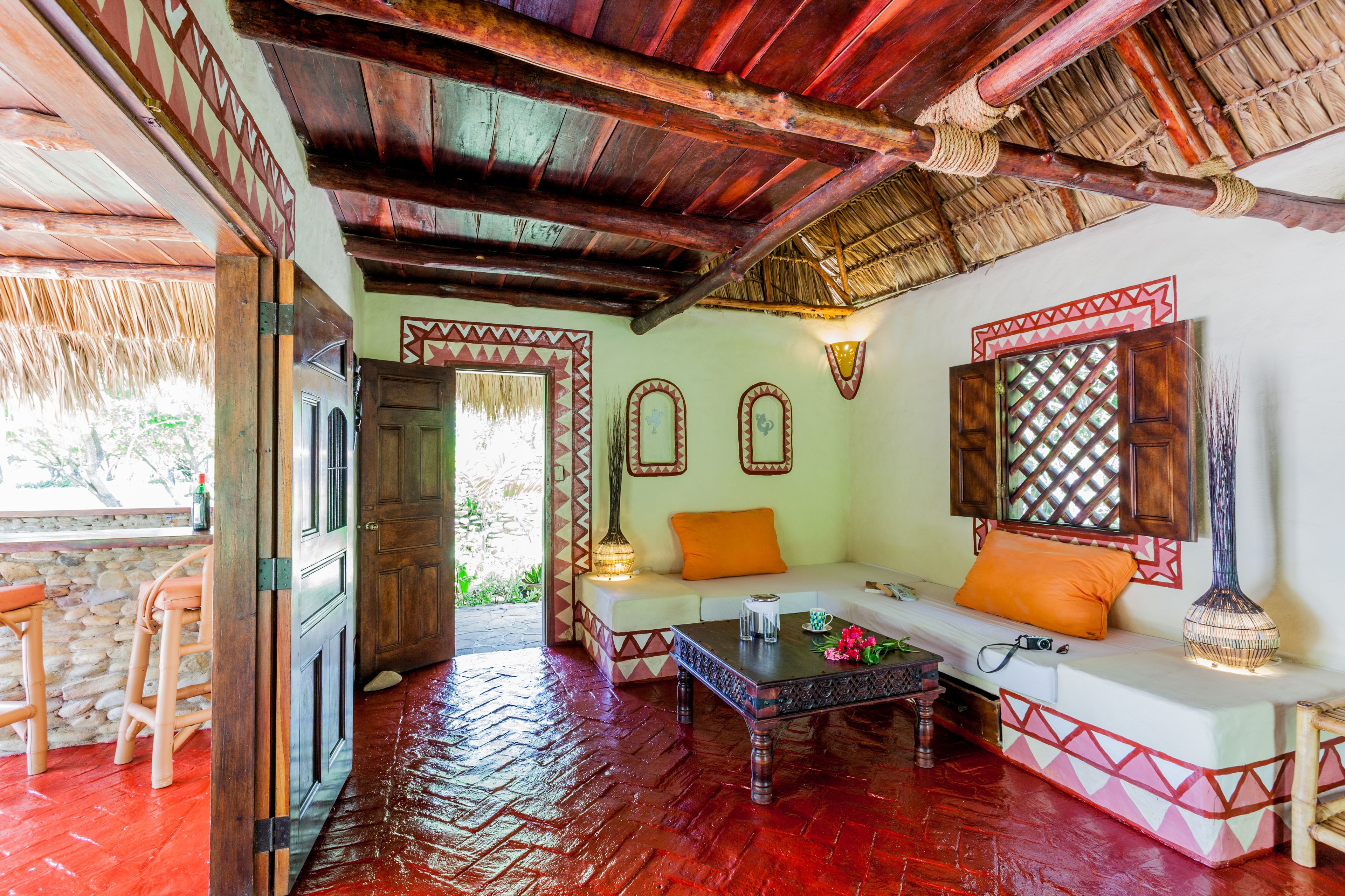 A traditional room with a wooden ceiling, a thatched roof outside, a sofa with cushions, a table with a lamp, and a decorative wall panel.